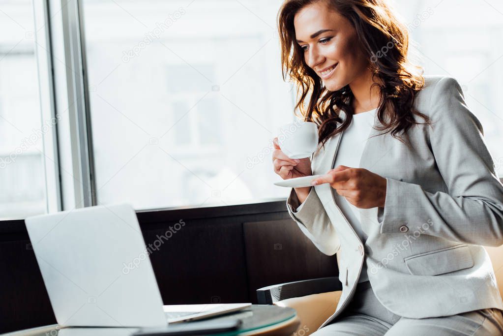 cheerful businesswoman using laptop while holding cup of coffee