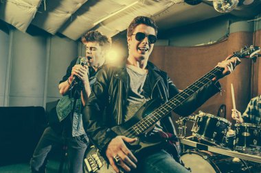 selective focus of guitarist in sunglasses playing electric guitar near rock band  clipart