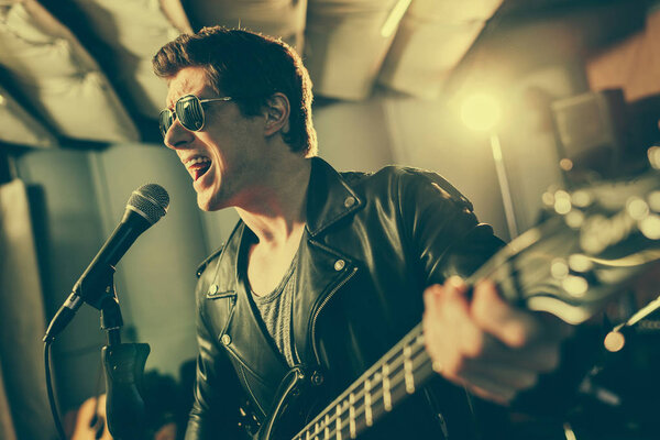 selective focus of guitarist in sunglasses singing song in microphone while playing electic guitar 