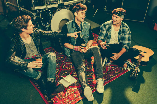 overhead view of cheerful rock band holding paper cups and talking while sitting on carpet  