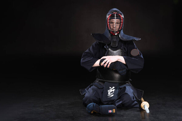 Kendo fighter in helmet sitting on floor and taking off glove on black