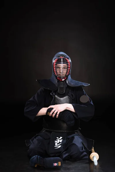 Kendo fighter in helmet sitting on floor and taking off glove on black