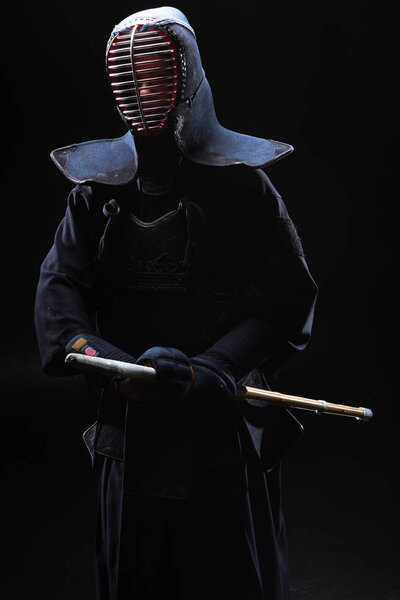 Kendo fighter in helmet holding bamboo sword on black