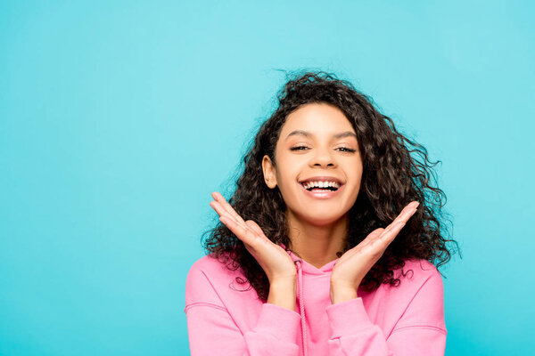 excited curly african american girl smiling while standing isolated on blue 