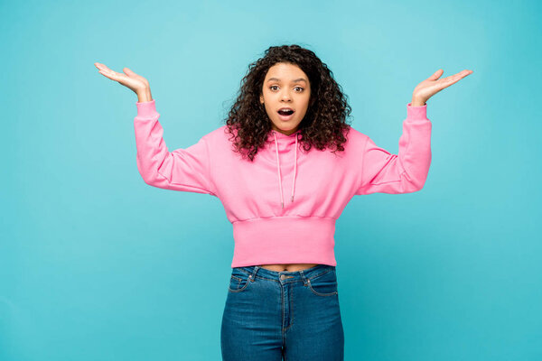 surprised curly african american woman showing shrug gesture isolated on blue 