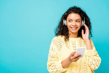 mavi üzerinde izole akıllı telefon tutarken neşeli kıvırcık Afrikalı Amerikan Kız kulaklıkta müzik dinleme