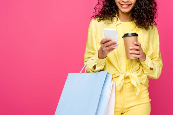 cropped view of african american woman with shopping bags holding smartphone and paper cup isolated on crimson