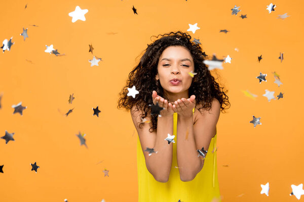 happy curly african american girl in yellow dress sending kiss near shiny confetti stars on orange 