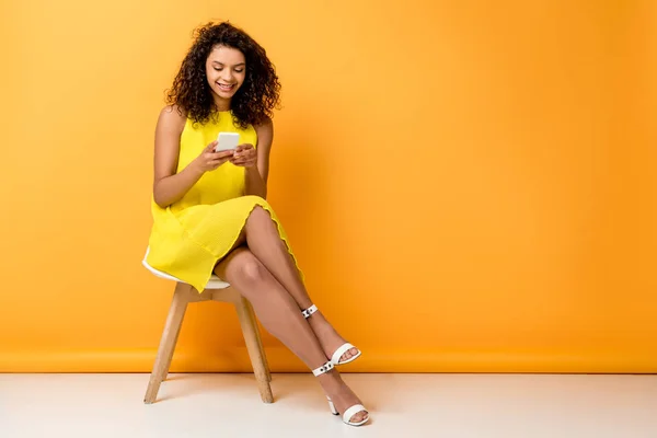幸せな巻き毛のアフリカ系アメリカ人の女性は椅子に黄色のドレスに座って オレンジ色のスマートフォンを使用します — ストック写真