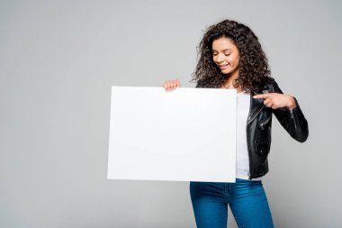 cheerful african american young woman pointing with finger at blank placard while standing isolated on grey clipart