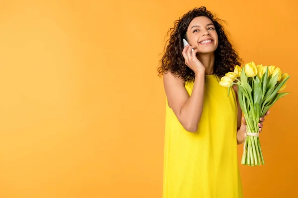 オレンジ色に孤立黄色のチューリップを保持しながら スマートフォンで話す幸せなカーリーアフリカ系アメリカ人の女性 — ストック写真