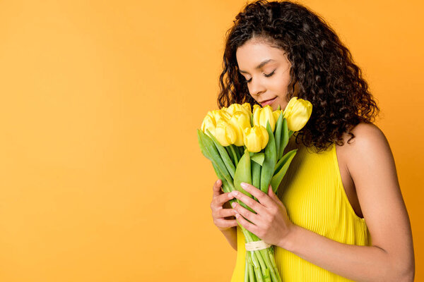 attractive curly african american woman smelling yellow tulips isolated on orange