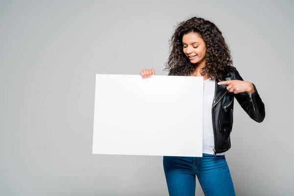 cheerful african american young woman pointing with finger at blank placard while standing isolated on grey