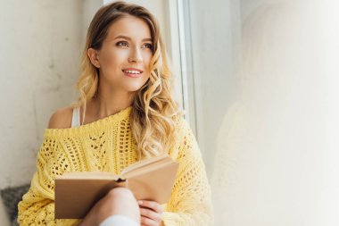 kopya alanı ile pencere eşik otururken güzel gülümseyen genç kadın tutmak kitap