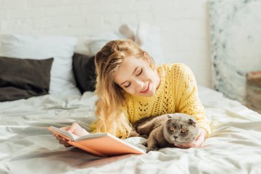 İskoç kat kedi ile yatakta yatarken güzel gülümseyen genç kadın kitap okuma 