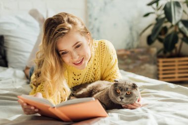 İskoç kat kedi ile yatakta yatarken güzel gülümseyen genç kadın kitap okuma 