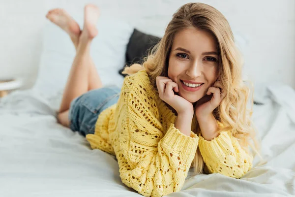 美丽的微笑的年轻女子在针织毛衣躺在床上 看着相机和手支撑脸 — 图库照片