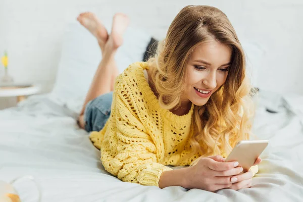 美丽的微笑的女孩在针织毛衣躺在床上 并使用智能手机 — 图库照片
