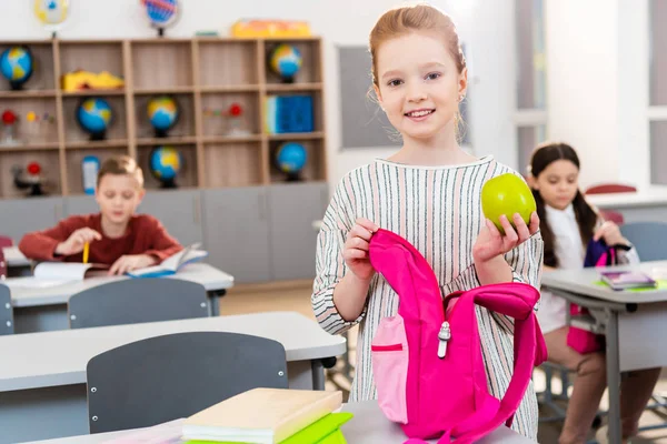 微笑的女学生拿着绿色苹果和粉红色的背包在教室里 — 图库照片
