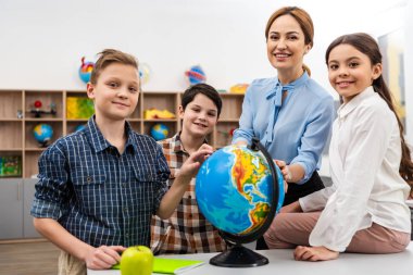 Teacher and pupils touching globe with smile while studying geography in classroom clipart