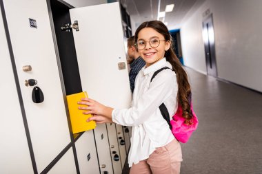 Joyful schoolgirl in glasses putting book in locker during brake in school clipart