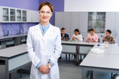 Smiling teacher in protective goggles standing in front of pupils during chemistry lesson