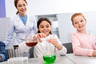 Smiling schoolgirl with beakers doing chemical experiment during chemistry lesson clipart