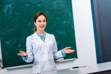 Beyaz ceket tahta önünde ders açıklayan gülümseyen kimya öğretmeni
