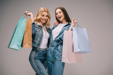 beautiful blonde and happy brunette women in denim clothes holding shopping bags and smiling   clipart