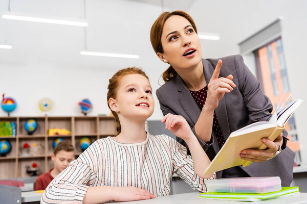 Teacher standing near desk and explaining lesson to pupil in classroom