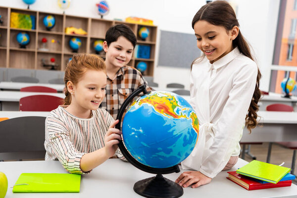 Three cheerful pupils playing with globe in classroom during geography lesson