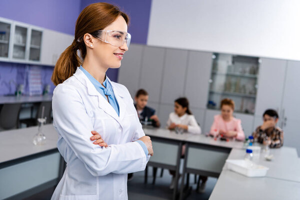 Smiling teacher in protective goggles standing with crossed arms in front of pupils during chemistry lesson