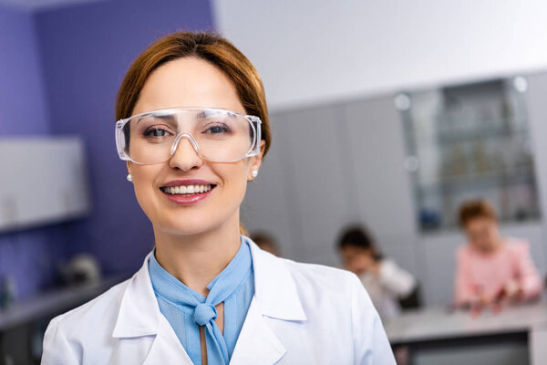 Smiling teacher in protective goggles standing in front of pupils during chemistry lesson