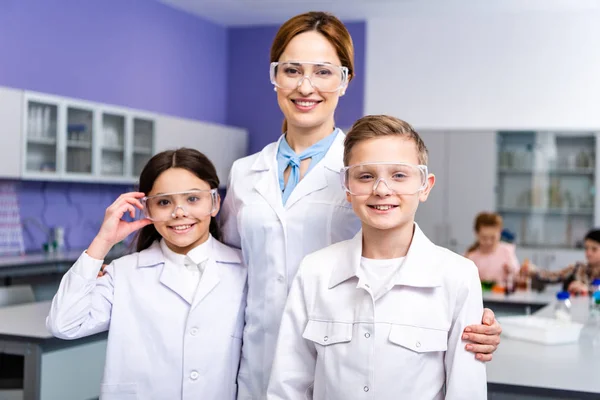 Smiling Teacher White Coat Protective Goggles Embracing Pupils Chemistry Lesson — Stock Photo, Image