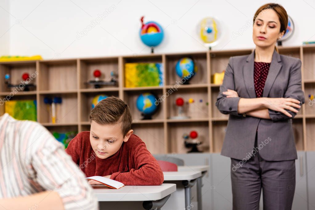 Serious teacher in suit standing with crossed arms and looking at pupils