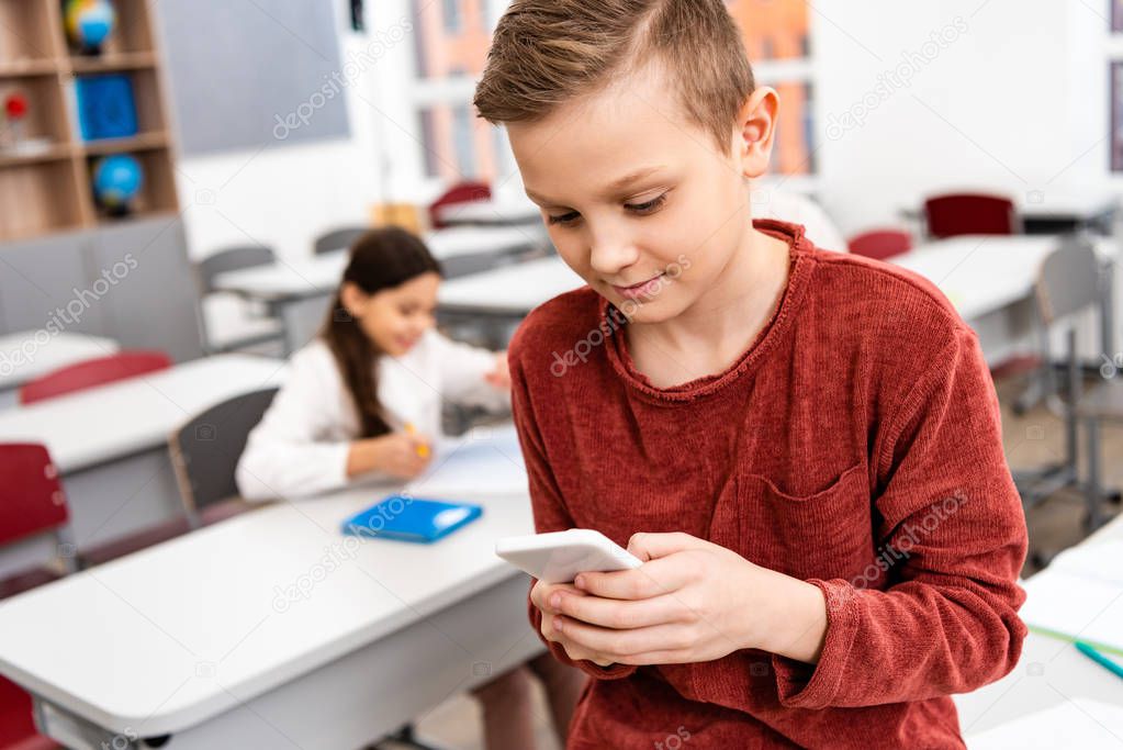 Interested schoolboy standing in classroom and using smartphone during brake 