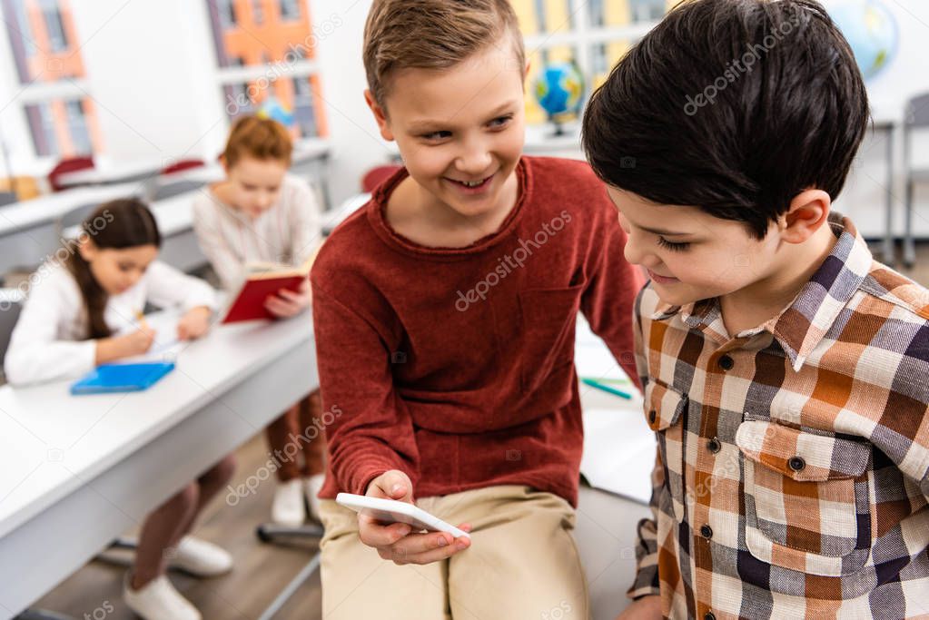 Two smiling schoolboys using smartphone in classroom during brake