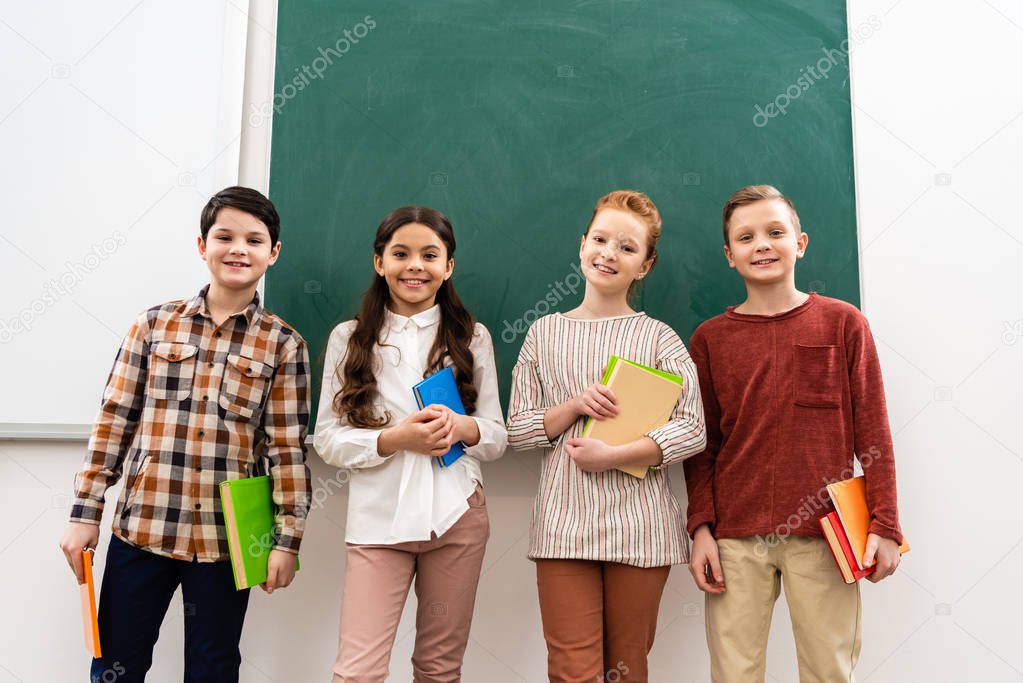 Smiling pupils with books and notebooks standing in front of blackboard