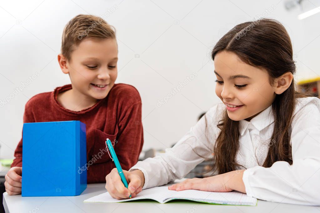 Smiling schoolgirl talking with friend while writing in notebook in classroom