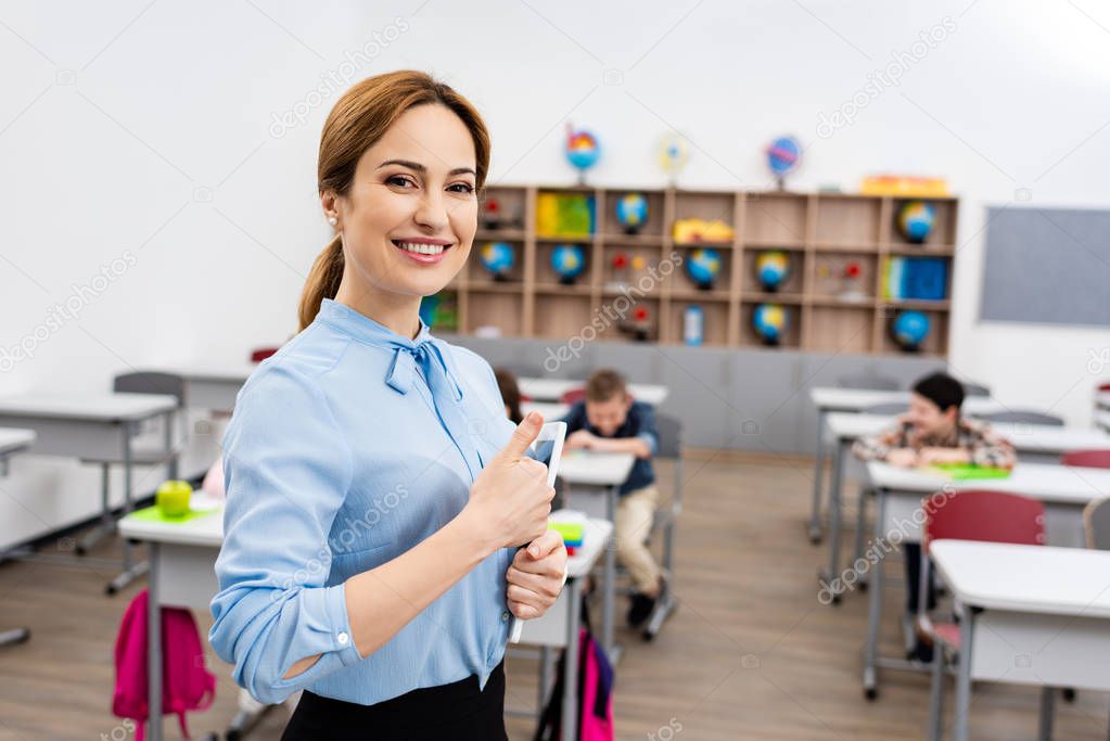 Laughing teacher in blue blouse standing in front of pupils and showing thumb up