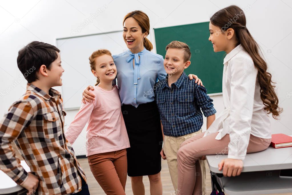 Laughing teacher embracing pupils in front of blackboard in classroom 