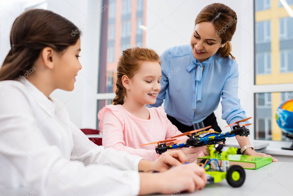 Smiling teacher standing near pupils with educational toys in classroom