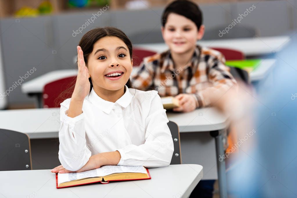 Excited schoolgirl with book raising hand up during lesson in classroom
