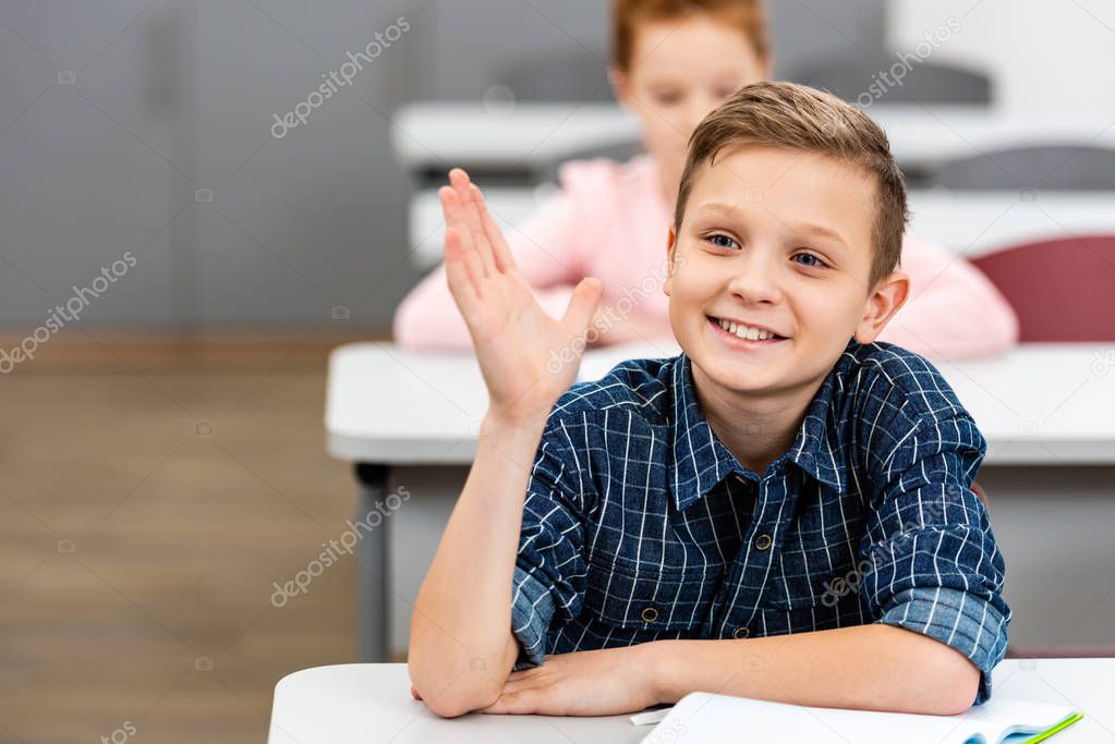 Schoolboy in checkered shirt raising hand up during lesson in classroom