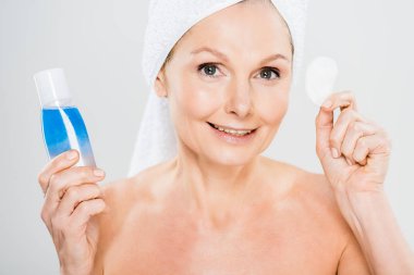 attractive and smiling mature woman in towel holding cotton pad and micellar water on grey background  clipart