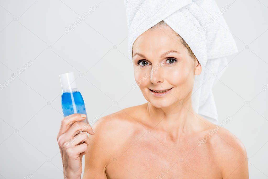 attractive and smiling mature woman in towel holding bottle with micellar water and looking at camera 