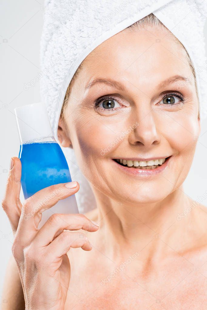 attractive and smiling mature woman in towel holding bottle with micellar water isolated on grey