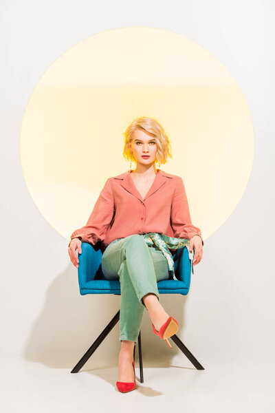 beautiful stylish girl in colorful clothes looking at camera and sitting in armchair on white with yellow circle