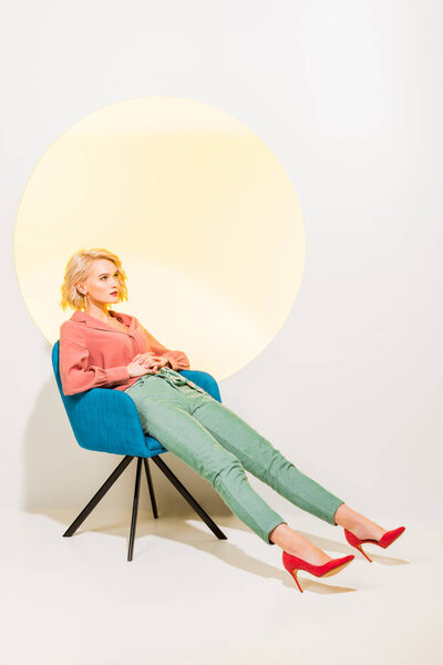 beautiful stylish girl in colorful clothes sitting in armchair on white with yellow circle and copy space