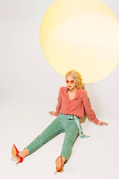 beautiful stylish girl in sunglasses sitting and posing on white with yellow circle and copy space